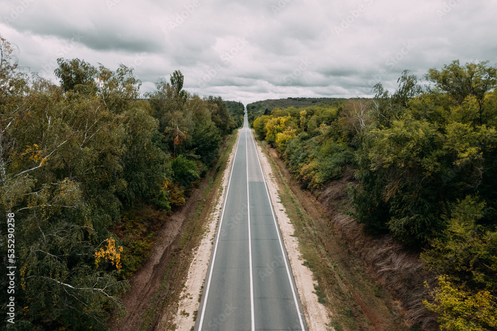 Aerial top view of the empty road between green and yellow autumn trees and cloudy sky. Drone shot of an asphalt road in the countryside.