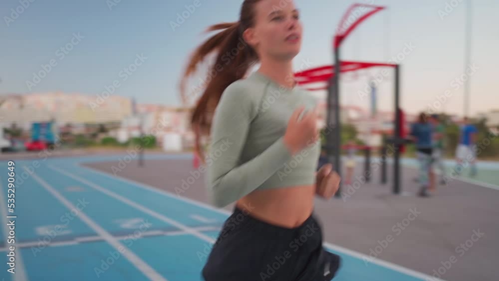 Female athlete running on track near sports ground. Fit caucasian young woman jogging evening dusk on running track. Athletic female jogger training outdoors. Healthy lifestyle concept Female athlete running on track near sports ground. Fit caucasian young woman jogging evening dusk on running track. Athletic female jogger training outdoors. Healthy lifestyle concept