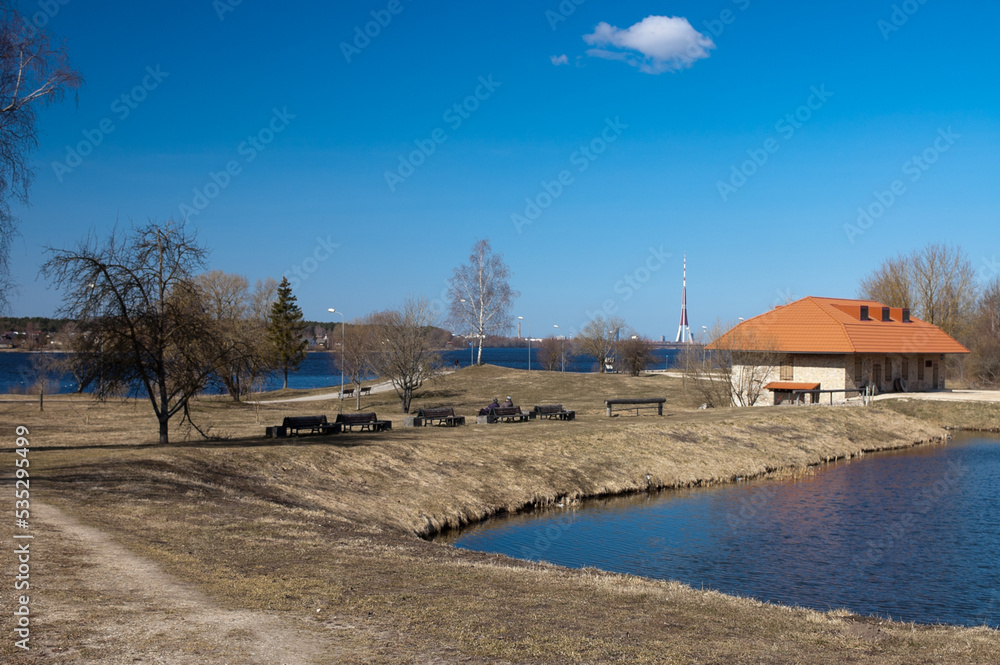 a park by the river, in the photo a pond, an old brick house, a river and a blue sky