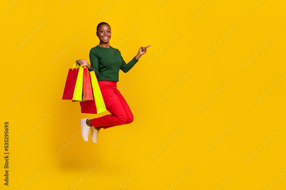 Full length photo of funny cute woman wear green shirt holding shoppers jumping empty space isolated yellow color background Full length photo of funny cute woman wear green shirt holding shoppers jumping empty space isolated yellow color background