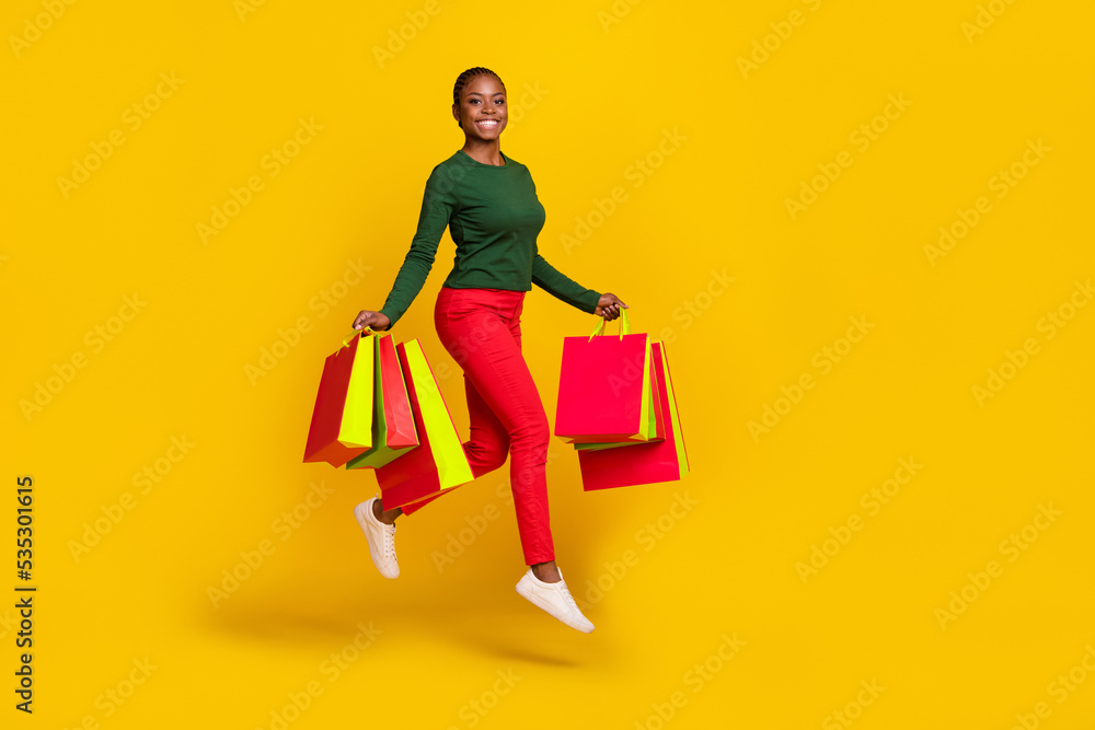 Full length photo of sweet funny woman wear green shirt holding shoppers jumping high isolated yellow color background Full length photo of sweet funny woman wear green shirt holding shoppers jumping high isolated yellow color background