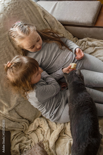 two little girls sisters sit at home on the couch and eat bread, their domestic gray cat is with them