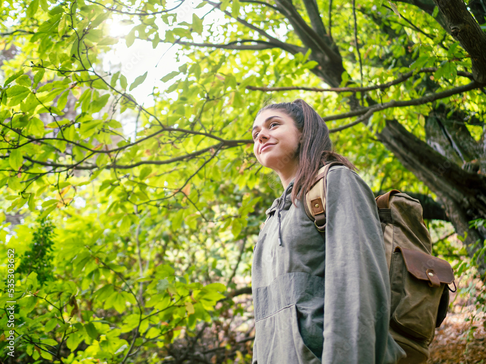 Side view of woman with backpack in the forest admiring the beauty of wild nature, looking up with a happy and satisfied smile. Side view of woman with backpack in the forest admiring the beauty of wild nature, looking up with a happy and satisfied smile.