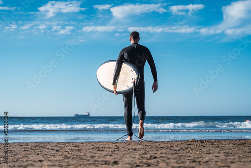 Obraz na płótnie Young man walks along the sand of the beach to enter the sea and do his favorite sport. The surf w salonie