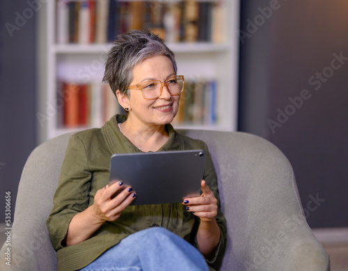 Mature woman using tablet computer while relaxing on sofa at home. Grey haired attractive woman holds digital tablet in hands sitting in chair.
