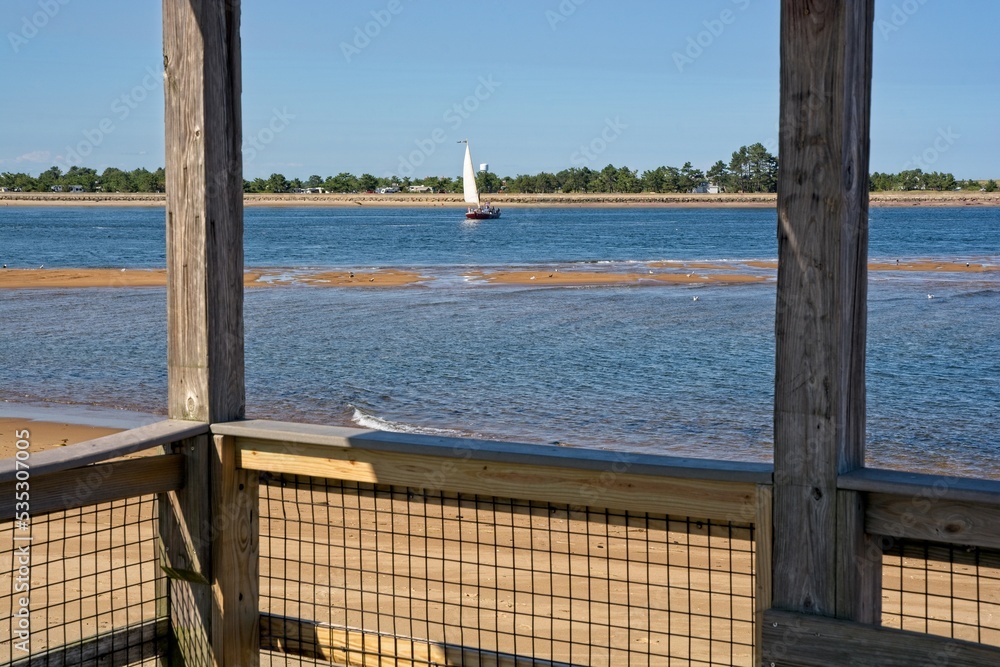 Fototapeta premium Sailboat tacking across Newburyport harbor channel viewed from boardwalk