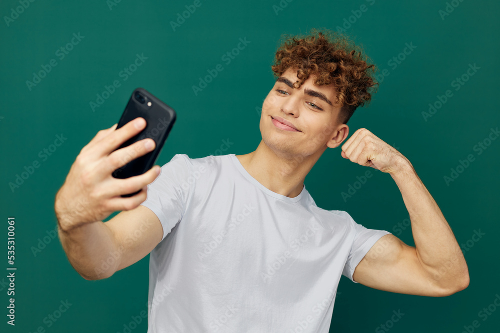 a joyful, cute man with curly hair stands against a green background and holds his black smartphone in his hand, photographing his biceps with a selfie camera a joyful, cute man with curly hair stands against a green background and holds his black smartphone in his hand, photographing his biceps with a selfie camera