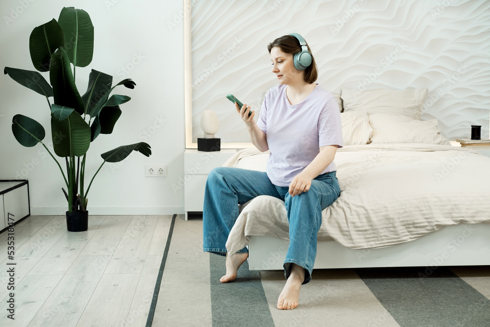 Smiling adult woman sitting on bed in bedroom wearing headphones and listening to music Smiling adult woman sitting on bed in bedroom wearing headphones and listening to music