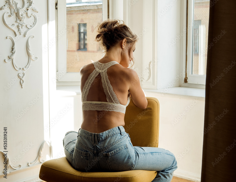 A beautiful young blonde woman in jeans and a bra sits in an armchair looking out the window
