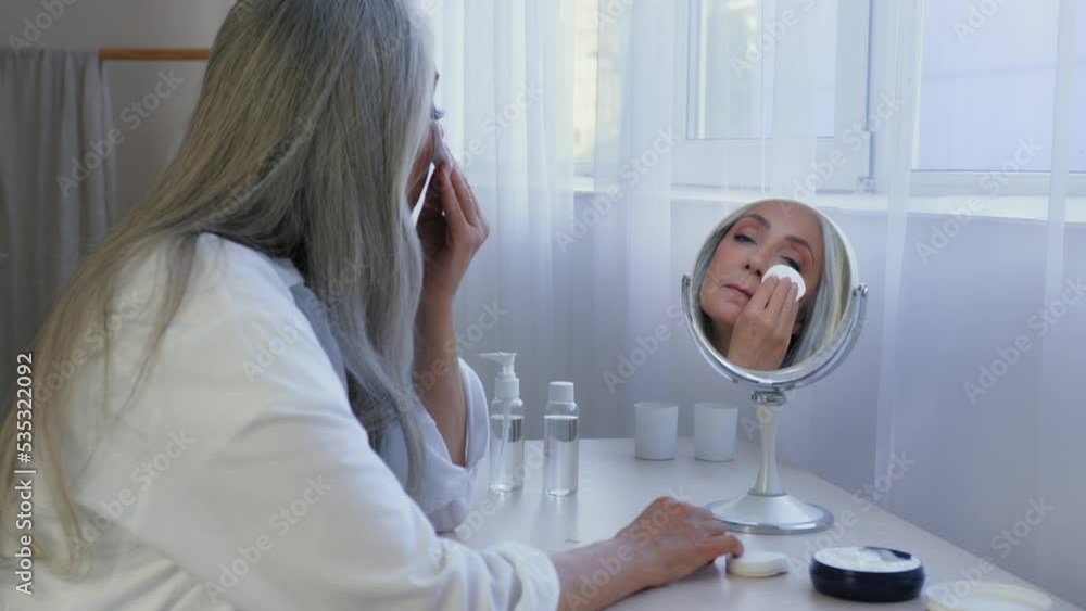 Gray-haired Caucasian mature middle-aged lady female old 60s woman at home looking mirror wipes face with cotton pad with tonic lotion serum smears face with cream removes makeup with sponge skincare Gray-haired Caucasian mature middle-aged lady female old 60s woman at home looking mirror wipes face with cotton pad with tonic lotion serum smears face with cream removes makeup with sponge skincare