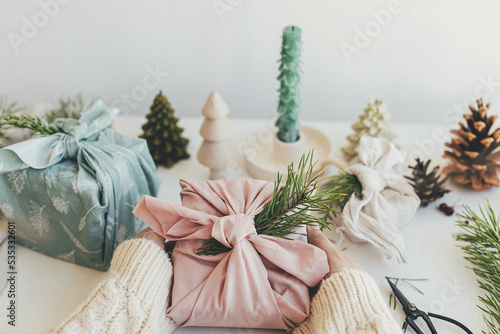 Furoshiki gift wrapping. Hands holding gift box wrapped in pink fabric with fir branch on white wooden table with eco friendly decor and candle. Zero waste Christmas concept. Merry Christmas!