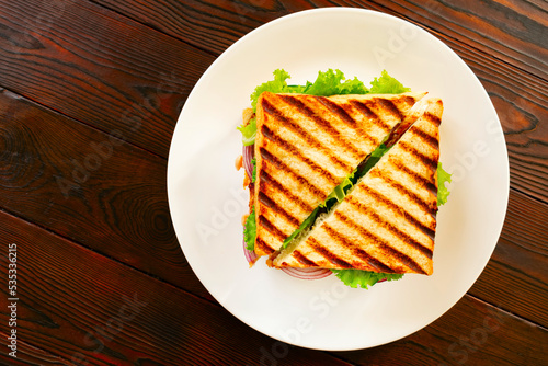Homemade club sandwich with toasted bread slices, chicken breasts, tomato, salad leafs, cucumber, onion on white plate. On wooden tabletop. Flat lay. 
