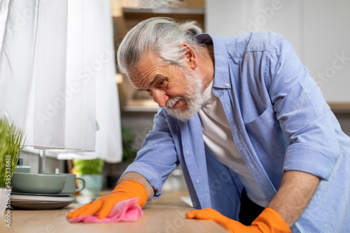 OCD Concept. Tidy Senior Man Cleaning Table In Kitchen With Rag photo