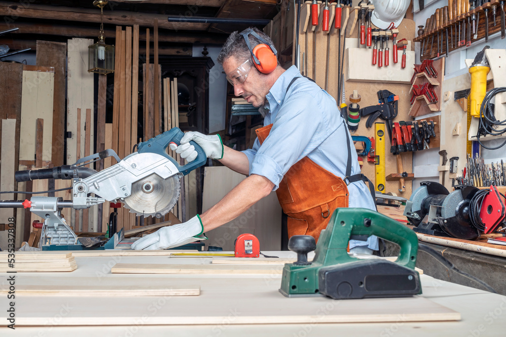 CARPENTER IN WORKSHOP WORKING WOOD WITH A MITER SAW. DIY AND PPE CONCEPT. PERSONAL PROTECTION EQUIPMENT FOR SAFETY AT WORK.