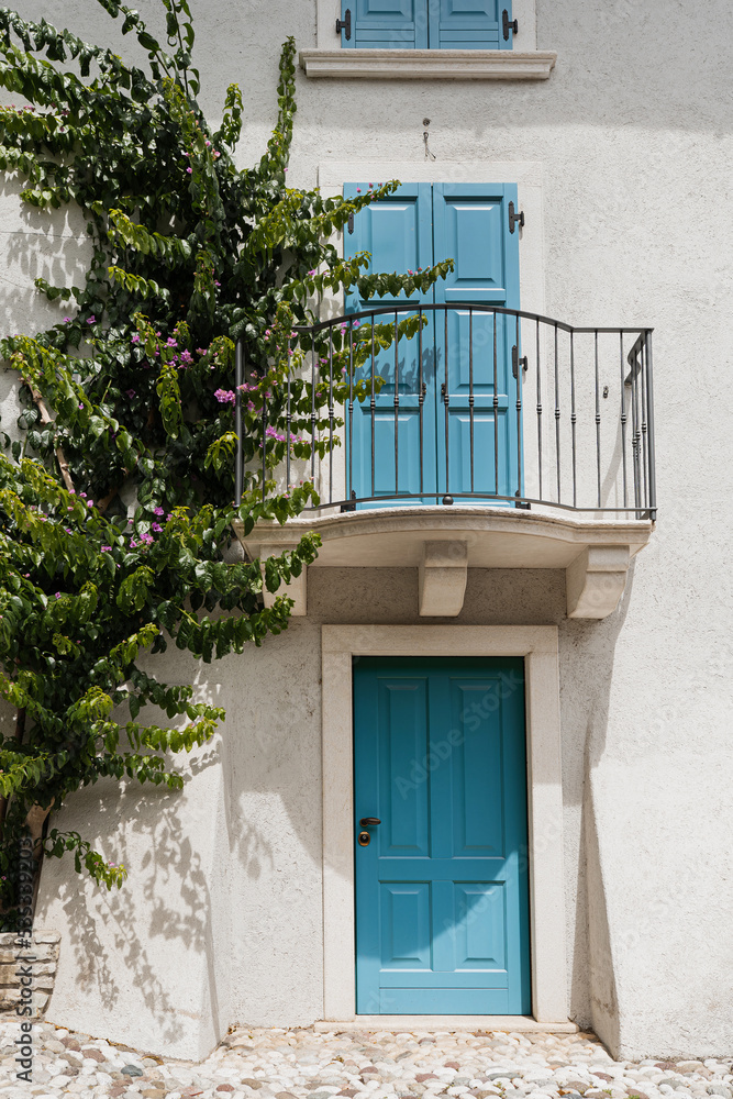 Old historic Italian architecture. Blue wooden door, balcony, blooming bush, white walls with sunlight shadows