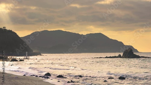 Video of Sakurai Futamigaura's Meoto Iwa Couple Rock and torii gate lighted by sunset light filtering through the clouds with the silhouette of people sightseeing on the Itoshima Beach of Fukuoka. photo