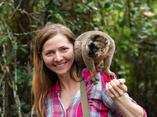 Young woman in shirt posing with small lemur on her shoulder, smiling, blurred jungle trees background