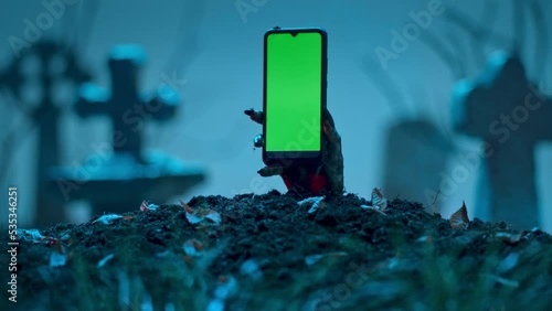 Zombie hand holds smartphone with green screen out of grave. Holiday event halloween concept. photo