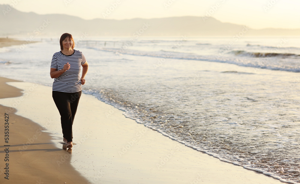Smiling middle aged woman running on the beach on sunrise. 40s or 50s attractive mature lady in sports clothes doing jogging workout enjoying fitness and healthy lifestyle at beautiful sea landscape.