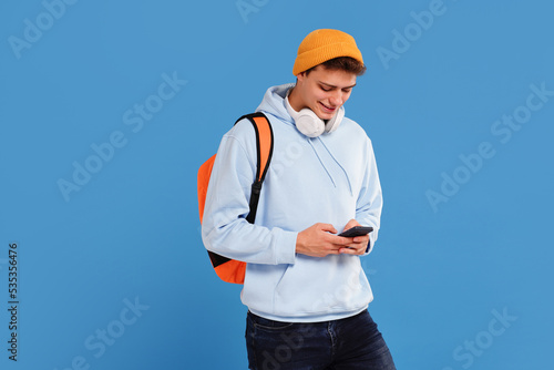 Young fashionable student guy with cap and backpack smiling, using mobile phone, isolated over blue studio background