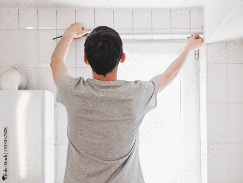 A young caucasian guy measures the width of a small window in the bathroom with a tape