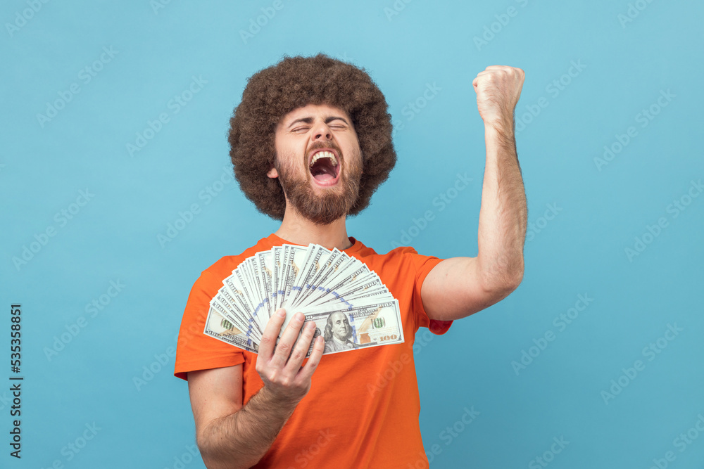 Portrait of extremely happy satisfied man with Afro hairstyle wearing orange T-shirt holding dollar bills and clenched fist, received an inheritance. Indoor studio shot isolated on blue background. Portrait of extremely happy satisfied man with Afro hairstyle wearing orange T-shirt holding dollar bills and clenched fist, received an inheritance. Indoor studio shot isolated on blue background.