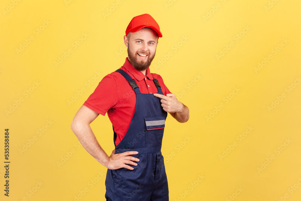 Side view of proud successful worker man standing and pointing at himself, bragging about the result of his work, wearing overalls and red cap. Indoor studio shot isolated on yellow background.