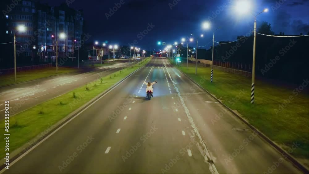 A drunk girl with a bottle of champagne in her hand rides a motorcycle through a deserted city at night. Drone shot. A drunk girl with a bottle of champagne in her hand rides a motorcycle through a deserted city at night. Drone shot.