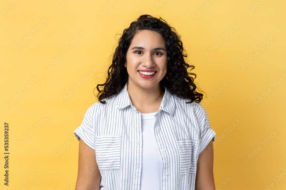 Portrait of cheerful positive woman with dark wavy hair looking at camera with toothy smile, expressing happiness, being in good mood. Indoor studio shot isolated on yellow background. Portrait of cheerful positive woman with dark wavy hair looking at camera with toothy smile, expressing happiness, being in good mood. Indoor studio shot isolated on yellow background.