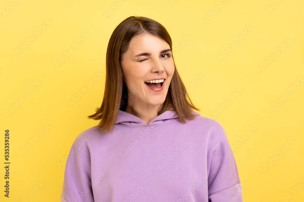 Portrait of positive optimistic woman standing and winking playfully, positive expression, looking at camera, wearing purple hoodie. Indoor studio shot isolated on yellow background. Portrait of positive optimistic woman standing and winking playfully, positive expression, looking at camera, wearing purple hoodie. Indoor studio shot isolated on yellow background.