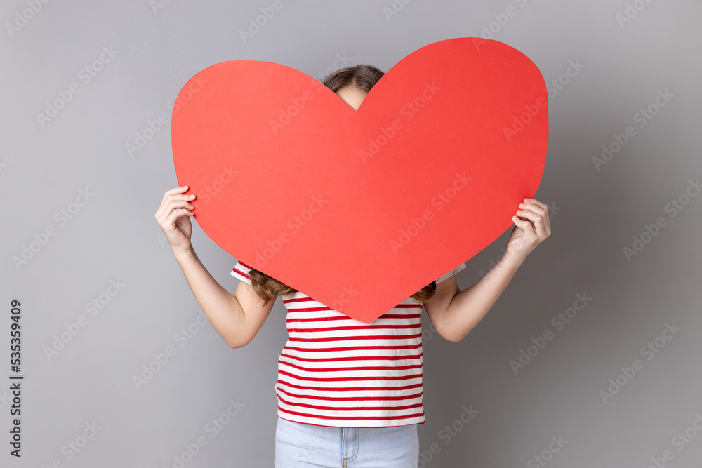 Portrait of anonymous unknown little girl wearing striped T-shirt hiding her face behind big red heart, hesitates to confess his feelings. Indoor studio shot isolated on gray background. Portrait of anonymous unknown little girl wearing striped T-shirt hiding her face behind big red heart, hesitates to confess his feelings. Indoor studio shot isolated on gray background.
