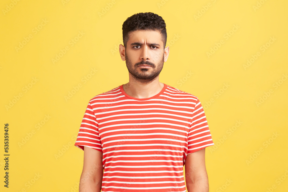 Portrait of serious sad upset man with beard wearing red T-shirt standing looking at camera, expressing sorrow and sadness, hearing bad news. Indoor studio shot isolated on yellow background. Portrait of serious sad upset man with beard wearing red T-shirt standing looking at camera, expressing sorrow and sadness, hearing bad news. Indoor studio shot isolated on yellow background.