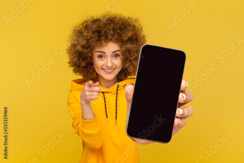 Φωτογραφία Portrait of attractive smiling woman with Afro hairstyle wearing hoodie showing big mobile display for copy space and pointing to camera, choosing you