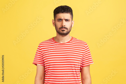 Portrait of serious sad upset man with beard wearing red T-shirt standing looking at camera, expressing sorrow and sadness, hearing bad news. Indoor studio shot isolated on yellow background.