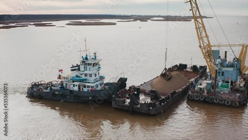 A floating barge with a pile of sand held by a tugboat. The bucket of the crane scoops the sand out of the pile. Aerial drone shot photo