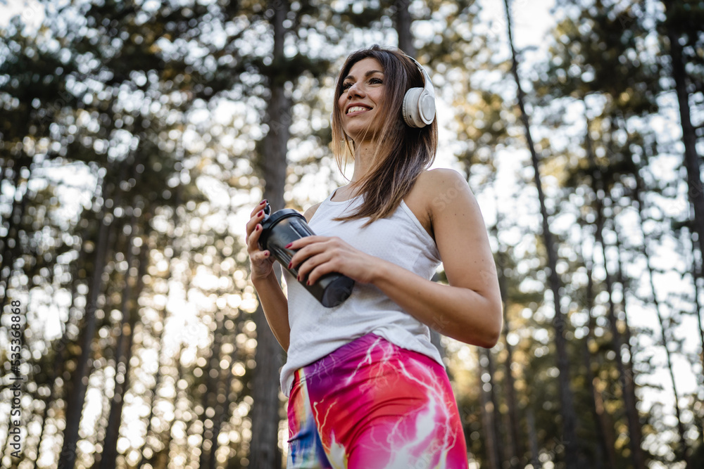 One woman happy caucasian female standing in the forest or park in woods with supplement shaker and headphones during training in nature taking a brake water drink and hydration real people copy space One woman happy caucasian female standing in the forest or park in woods with supplement shaker and headphones during training in nature taking a brake water drink and hydration real people copy space