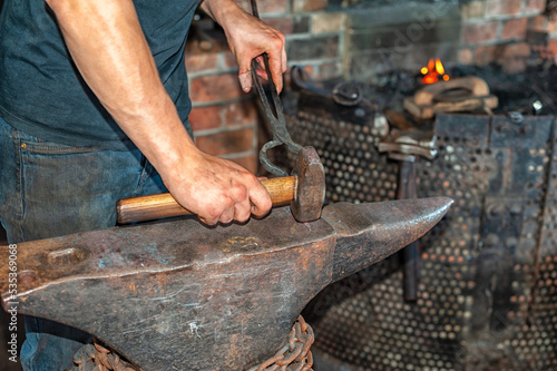 A forger wearing blue jeans and a t-shirt holds fire thongs and a metal twisted bolt on a vintage anvil. There's rounding hammer tool with a wooden handle in the blacksmith foundry shop with a furnace