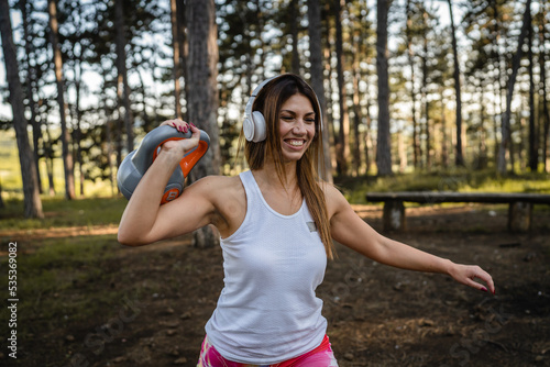 Fototapeta Naklejka Na Ścianę i Meble -  One woman adult caucasian female athlete using kettlebell girya weight during training in the forest park woods in summer day with headphones happy smile brunette health and fitness concept copy space