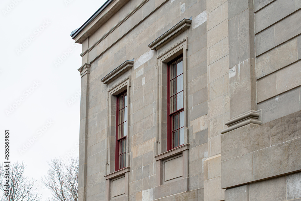 The exterior of a vintage limestone block wall with multiple windows. The tall government building has a blue sky with clouds in the background. The trim around the double hung windows is red wood