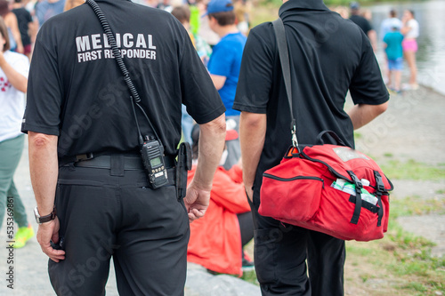 Medical first responders walking along a road wearing black uniforms, with medical first responder in grey letters across the back of the paramedic. The EMT is carrying a red first aid kit and radio.