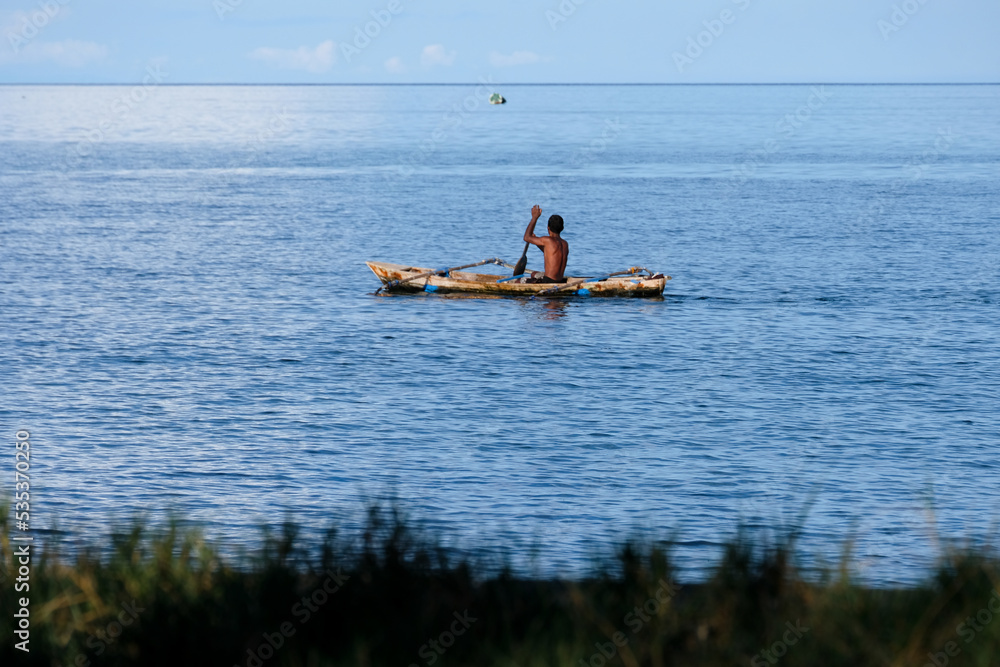 Naklejka premium A Timorese fisherman paddling a traditional wooden fishing canoe boat over calm placid ocean water on tropical island in Timor Leste, Southeast Asia
