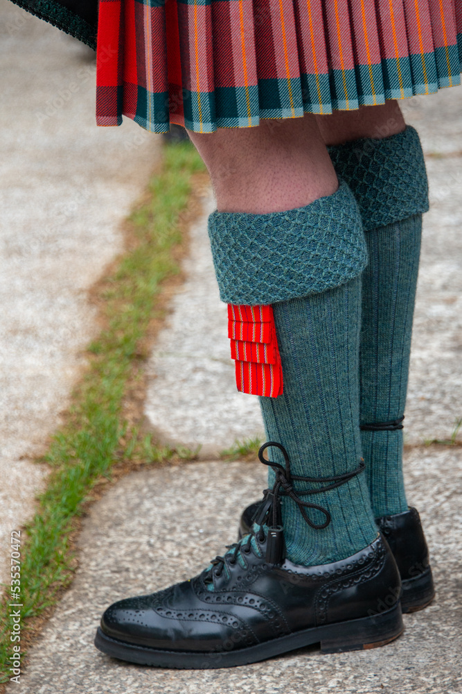 A male Irish piper stands on a sidewalk during a parade wearing a red, green, and white colored kilt. The man has solid green wool hoses with vibrant red tassels and shiny black dress shoes. 