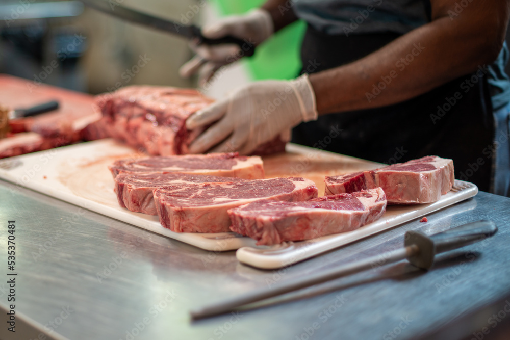 A close up of a large prime rib roast which is sitting on a brown plastic cutting board. The meat has some marbling and multiple ribs. The chef is using a long knife to cut steaks from the fresh meat.