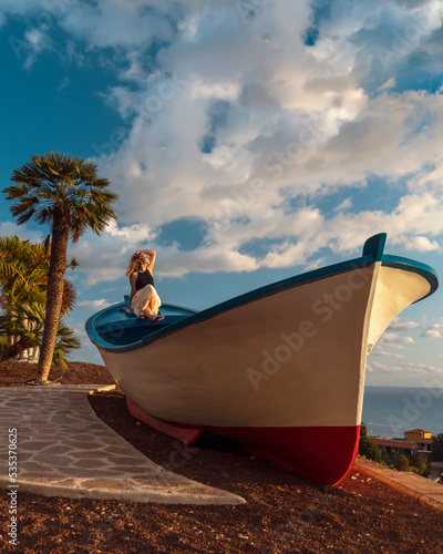 Girl sitting on the wooden fishing boat. Tropical holiday in Los Gigantes, Tenerife, Canary islands, Spain. Palm in the background. Beautiful colorful sky with sun and clouds.