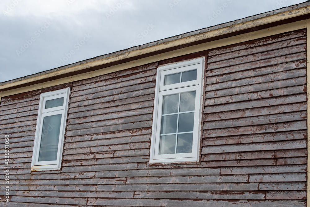 Two white vinyl windows with white trim and clear glass. The exterior wall has pine clapboard siding with red paint peeling. The boards are weathered and worn on the exterior of an old flat roof barn.