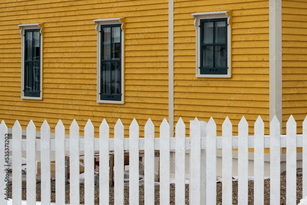 A white wooden picket fence in the foreground with a bright yellow cape cod siding covered building in the background. The house has multiple vintage windows in the exterior wall with green trim. 