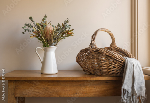 Australian native flowers in white jug wtih wicker basket and grey scarf on oak side table against beige wall