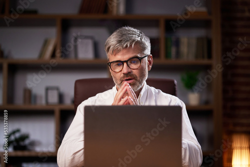 Gray-haired manager in glasses uses a laptop, conducts a video conference in the office. Middle aged businessman participating in online webinar looking at computer webcam while sitting at workplace.