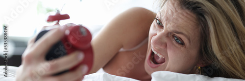 Woman lying in bed with alarm clock in her hands and screaming