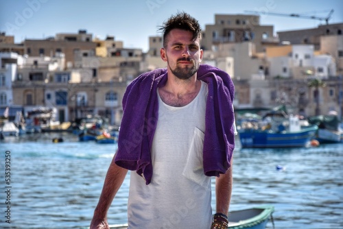 Mature man with purple towel in his shoulder standing by waterside against town and fishing boats photo
